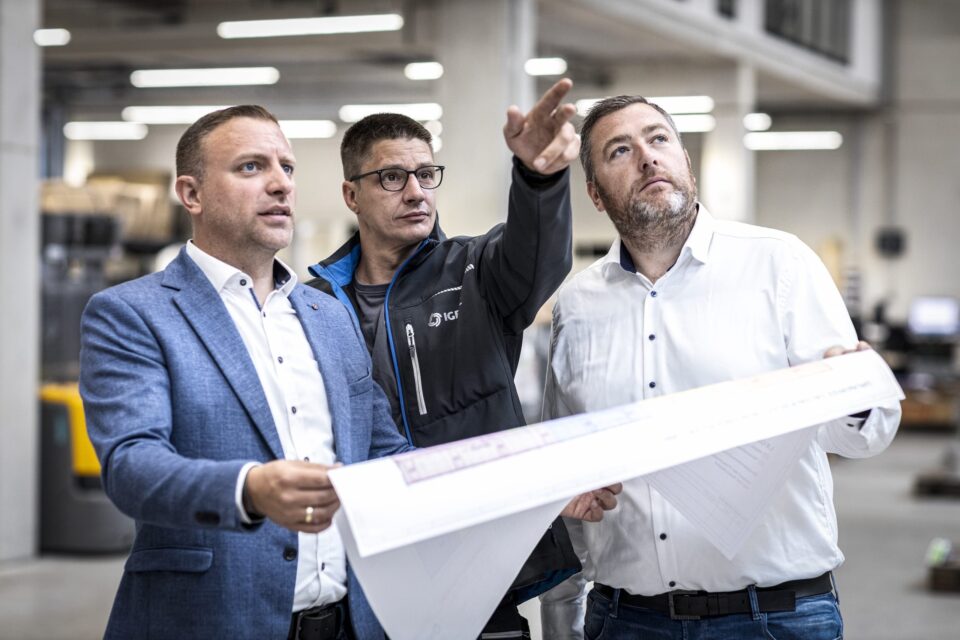 Drei Männer betrachten Baupläne in einer Fabrikhalle.