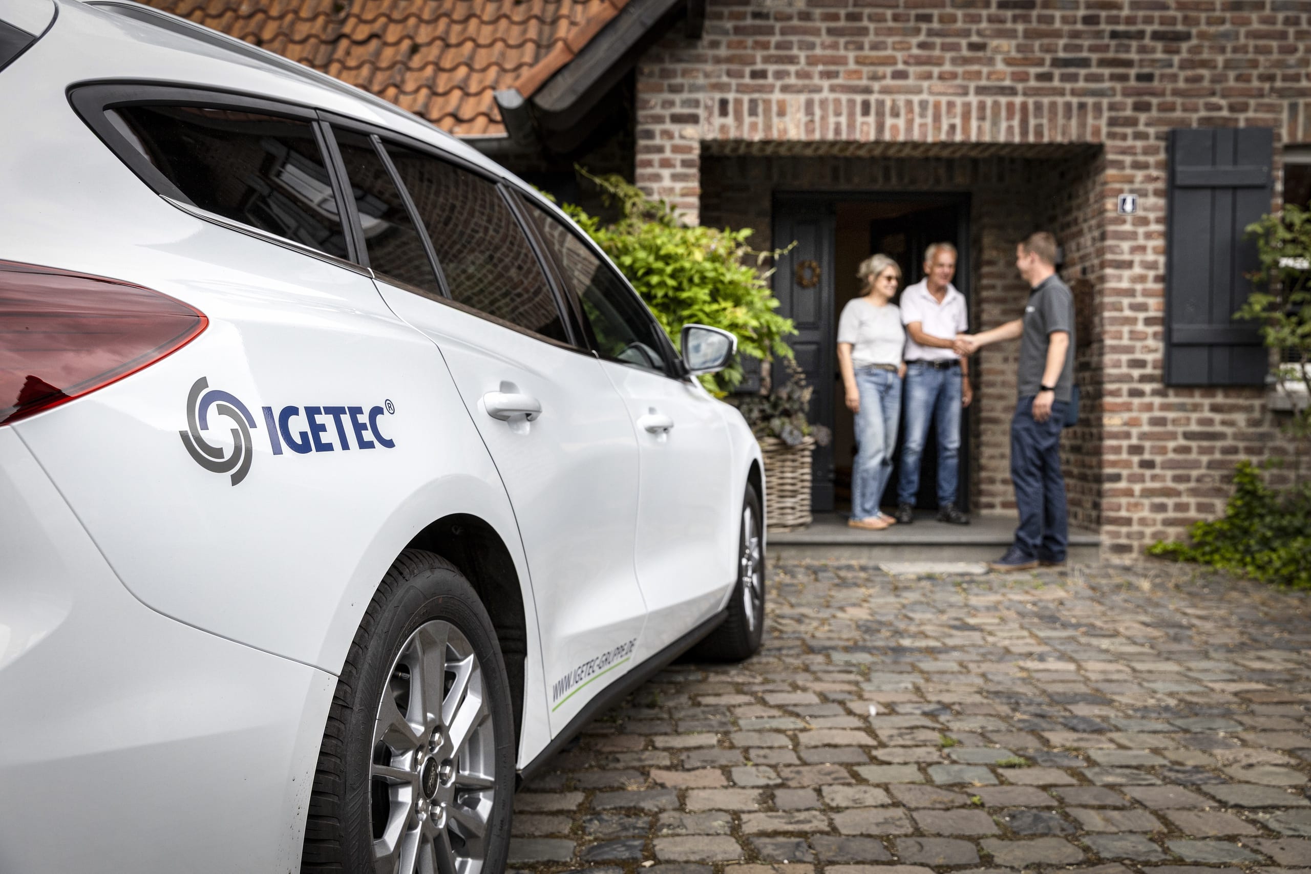IGETEC Auto vor Haus, Menschen begrüßen sich.