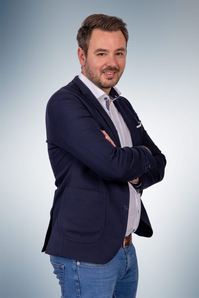 Mann im Anzug mit verschränkten Armen lächelt.