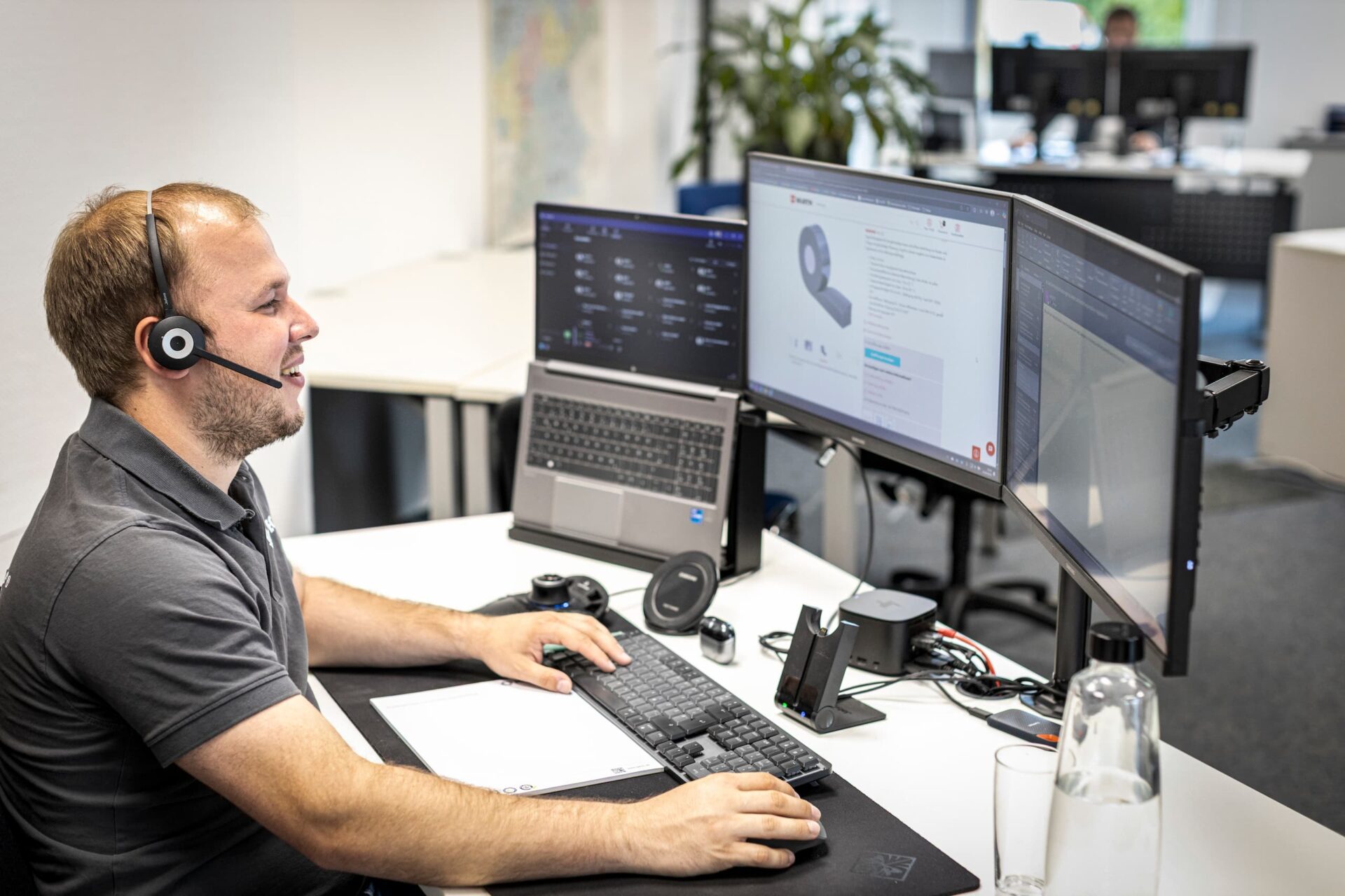 Mitarbeiter von IGETEC sitzt mit Headset am Schreibtisch vor zwei Monitoren und einem Laptop, arbeitet am Computer und telefoniert gleichzeitig im modernen Büro.