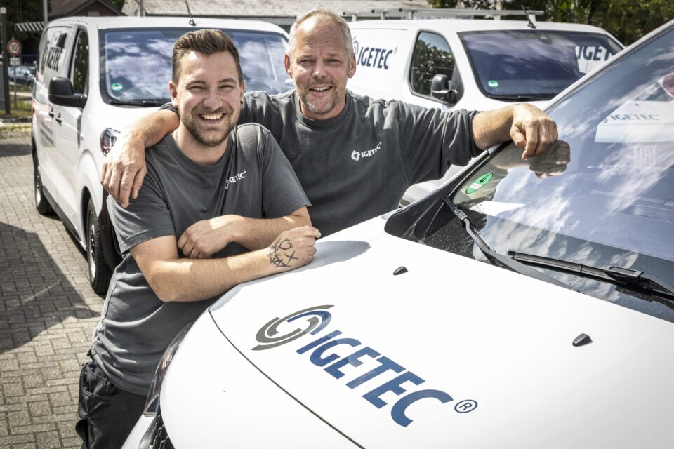 Zwei Männer vor IGETEC Firmenwagen lächelnd.